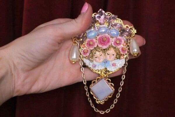 A hand holding a necklace with two angels and flowers.
