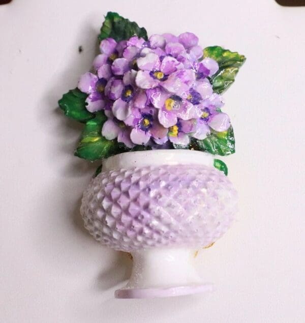 A purple flower in a white vase on the wall.