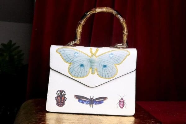 A white purse with butterflies and beetles on it.