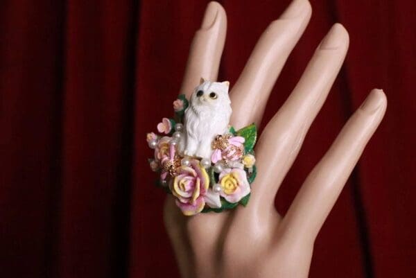 A hand wearing a ring with flowers and a cat on it.