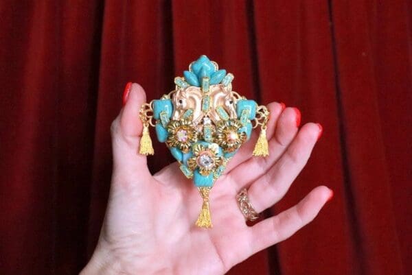 A hand holding a turquoise and gold brooch.