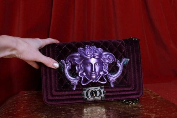 A woman holding onto a purple purse with an image of medusa.