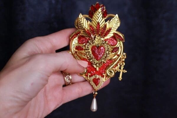 A hand holding a red and gold heart shaped brooch.