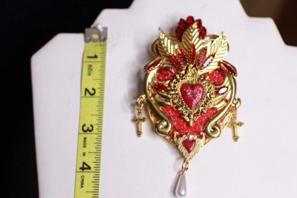 A large red and gold heart shaped brooch with a pearl dangling from it.