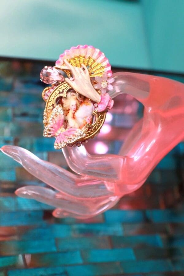 A pink hand holding a ring with a shell and angel on it.